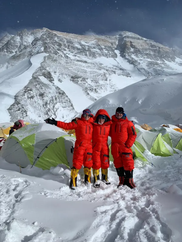 Three climbers in red down suits at a high camp on Everest's North Side, with the snow-covered upper mountain rising behind the tents.