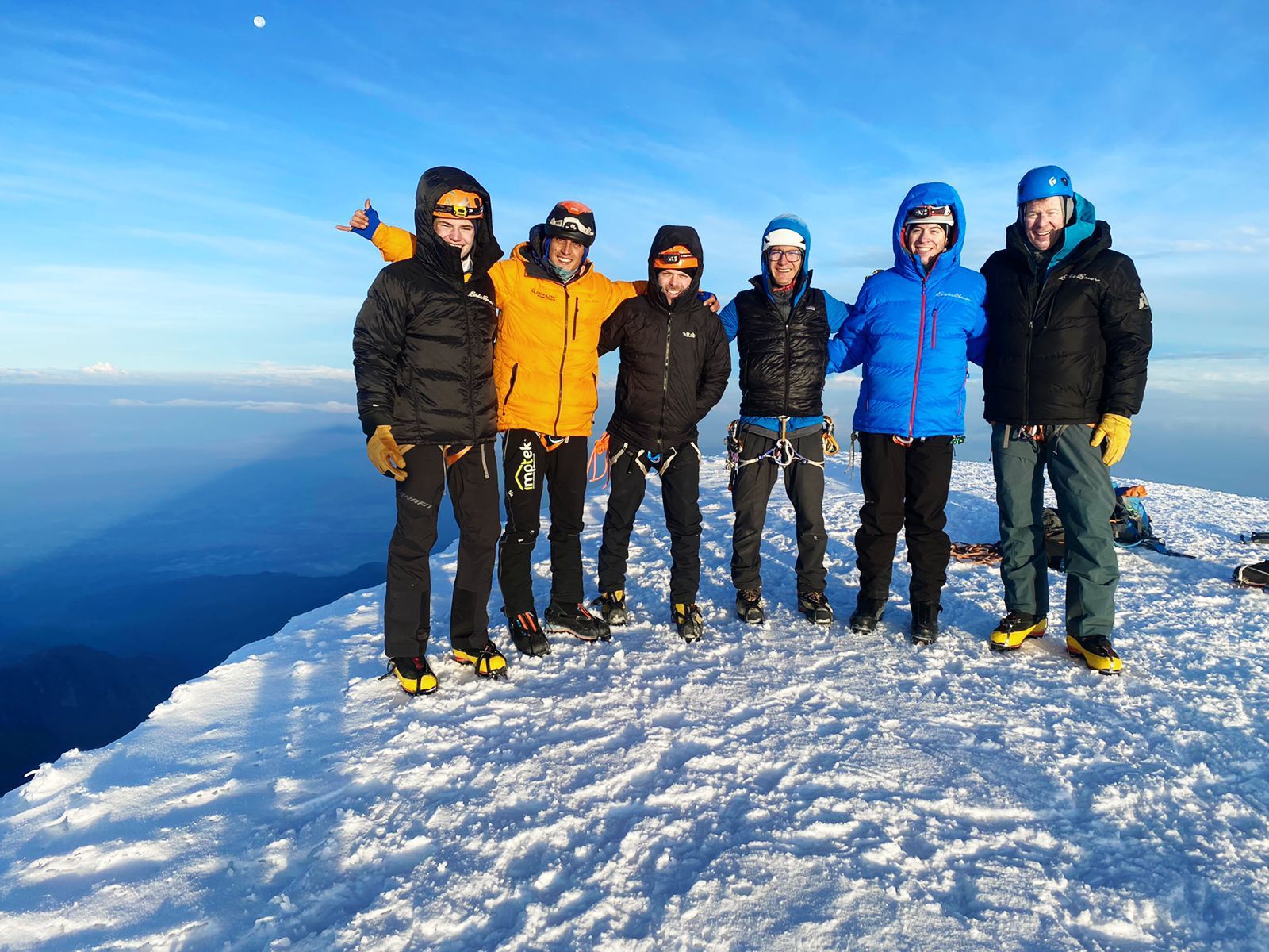 A group of 6 climbers stand atop a snowy mountain summit beneath a blue sky.