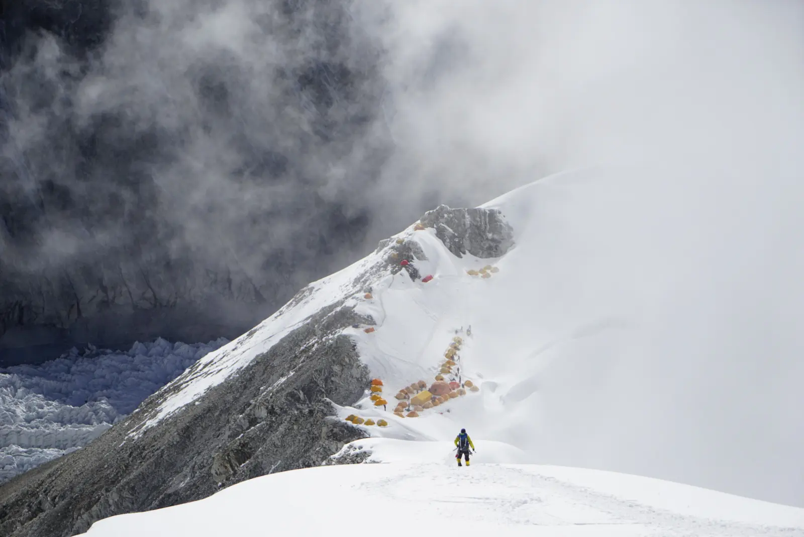 A climber moving up along a snowy ridge, with tents in the background on a Cho Oyu Expedition with Alpenglow Expeditions.