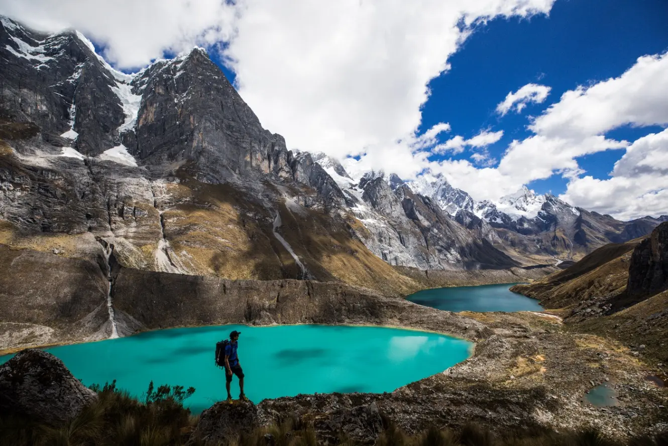 A hiker on the Cordillera Huayhuash Trek with a green lake and high peaks in the background