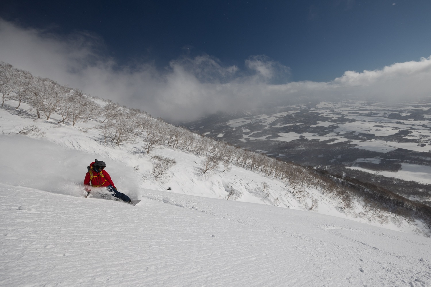 A backcountry snowboarder carves through powder on a white snowy hillside in Japan with a blue sky above.