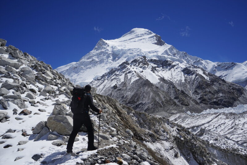 A climber stands on a mountain path with Cho Oyu in the background.