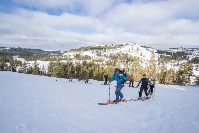 An Alpenglow Expeditions ski guide leads a group of intro to backcountry skiers in the Tahoe backcountry on snow with a mountain in the background, and a partly cloudy sky.