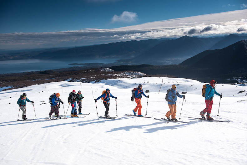 A line of skiers ascend a skin track in a wide open snow field on a Chilean volcanoe under blue skies and above a mountain lake.