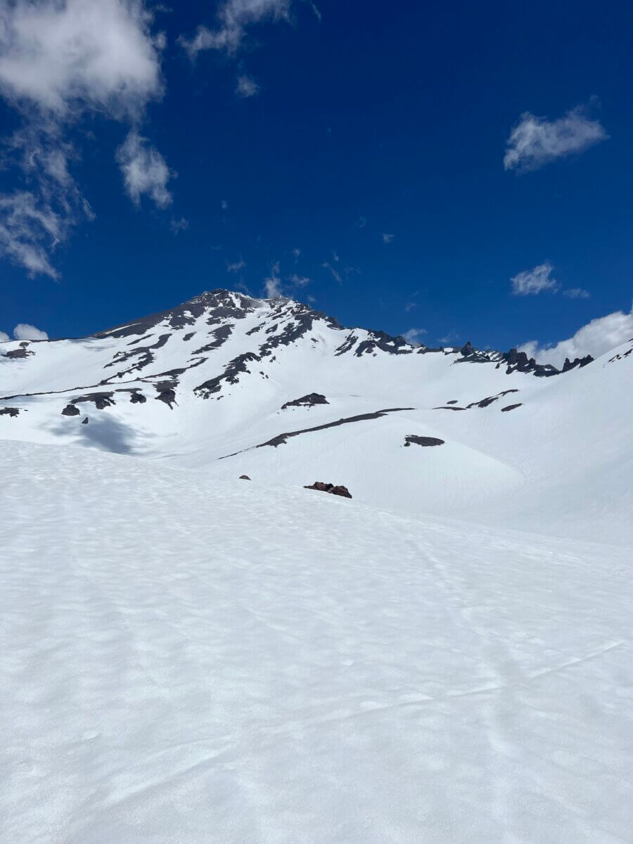 A view looking up Mount Shasta showing snow in the foreground, a rocky ridgeline and a beautiful blue sky with a few clouds
