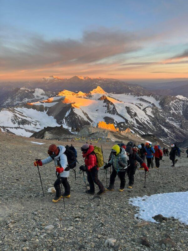 A group of climbers walks across a tallus field while climbing Aconcagua with the sun rising on the snowy mountains behind them.