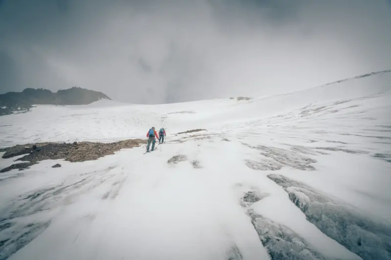 Skiers ascend a glacier through crevassed terrain under dark, cloudy skies.