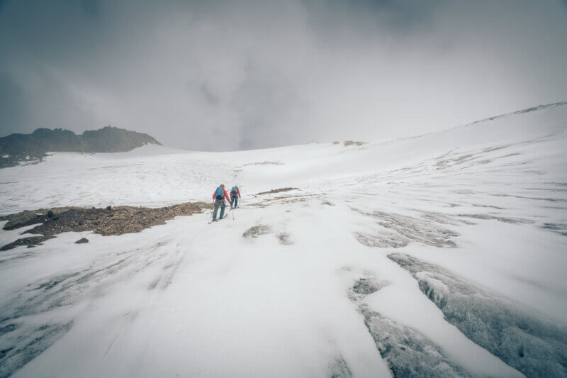 Skiers ascend a glacier through crevassed terrain under dark, cloudy skies.