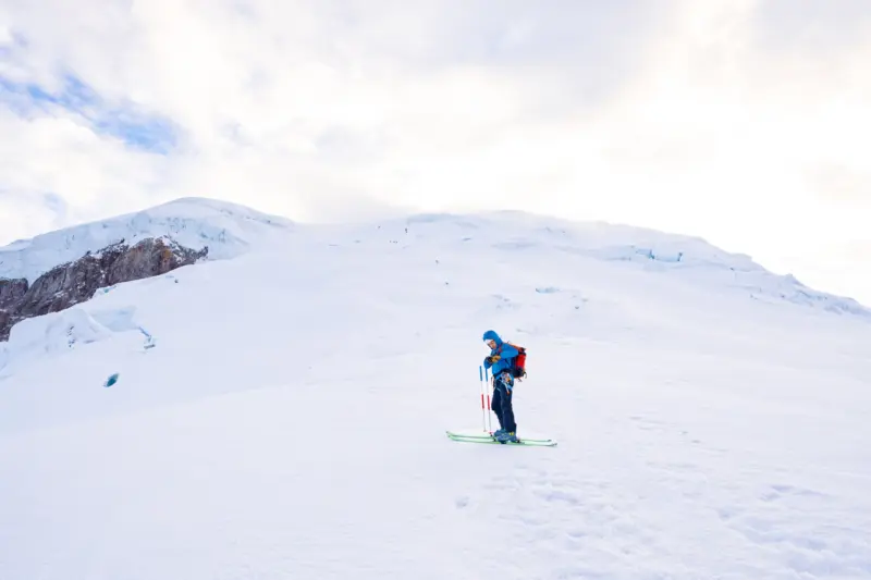A skier stands on a blank snowfield beneath the summit of Cayambe and cloudy skies above.