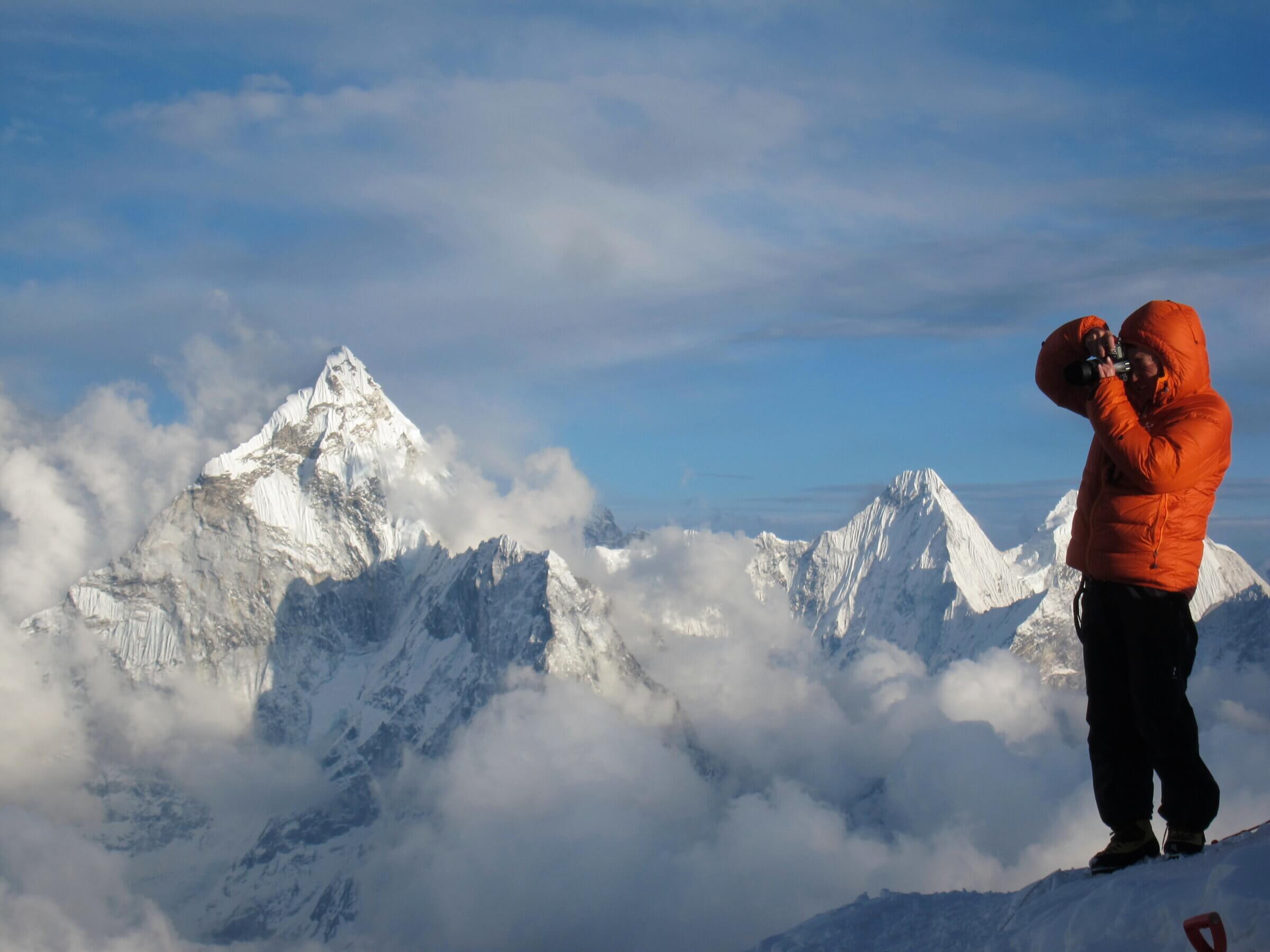 A photographer stands on a ridge with a large Himalayan mountain behind him.