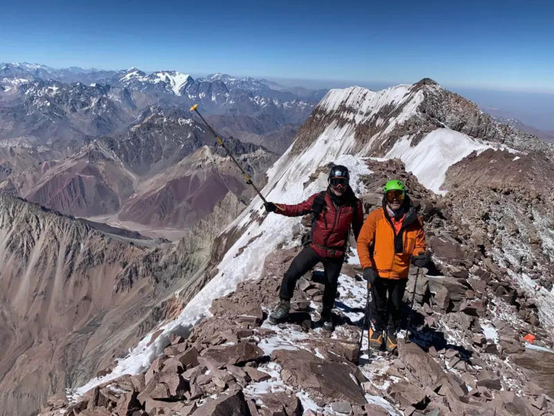Two climbers in purple and orange jackets stand on a rocky and snowy mountain ridge.