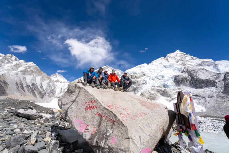 A group of five sits atop the rock at Everest Base Camp with the Himalaya in the background.