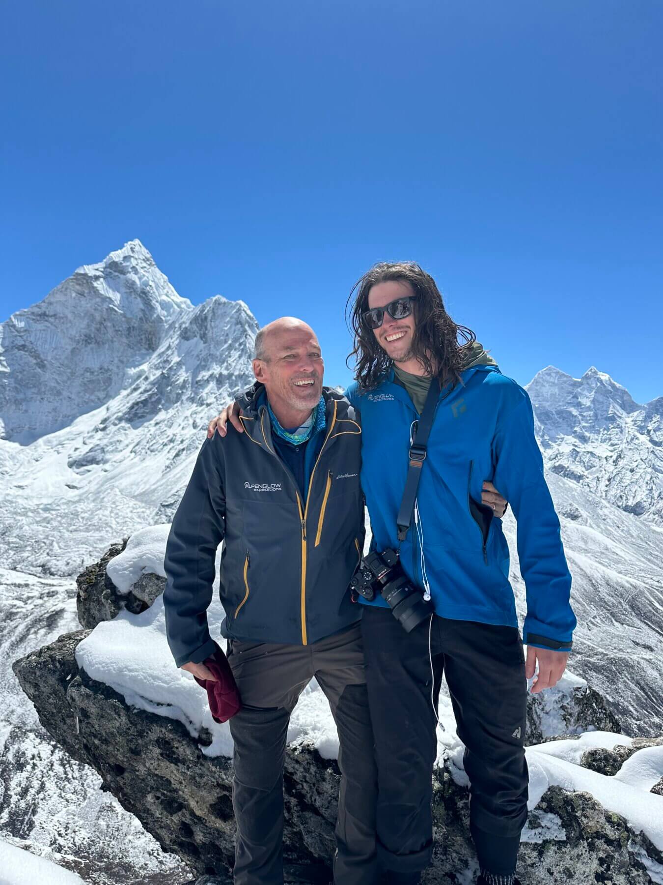 Two people stand on top of a mountain smiling with Ama Dablam in the background.