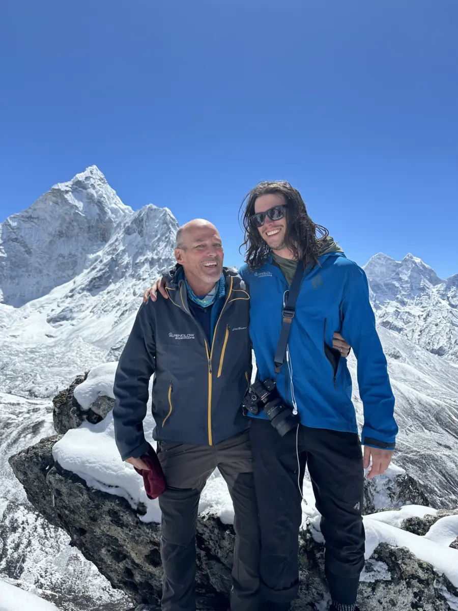 Two people stand on top of a mountain smiling with Ama Dablam in the background.