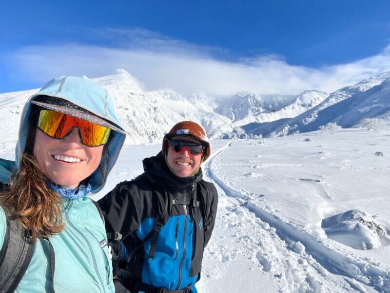 Two skiers take a selfie next to their skin track in a field of white snow in Japan beneath a big blue sky during a guided Japan ski trip with Alpenglow Expeditions.
