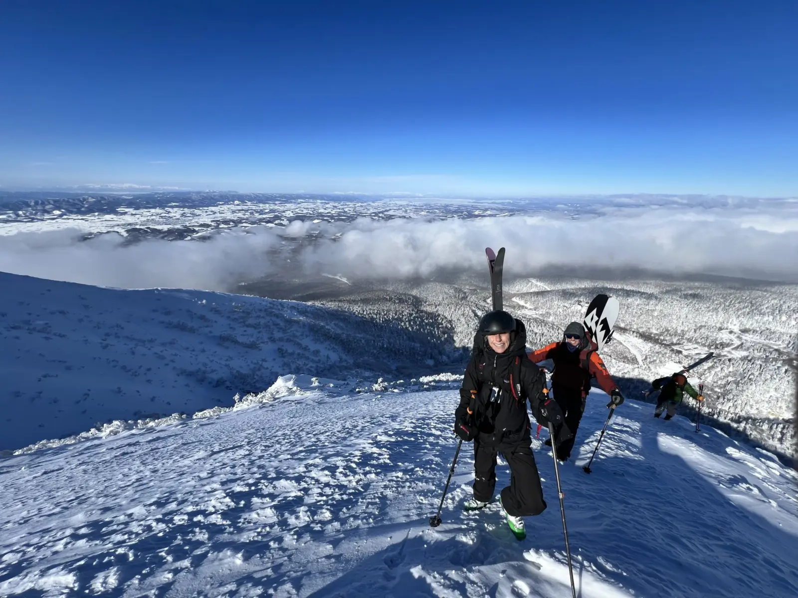 Two skiers stand with skis on their backs on a white snowy slope above the forest in Japan. The sky is blue.