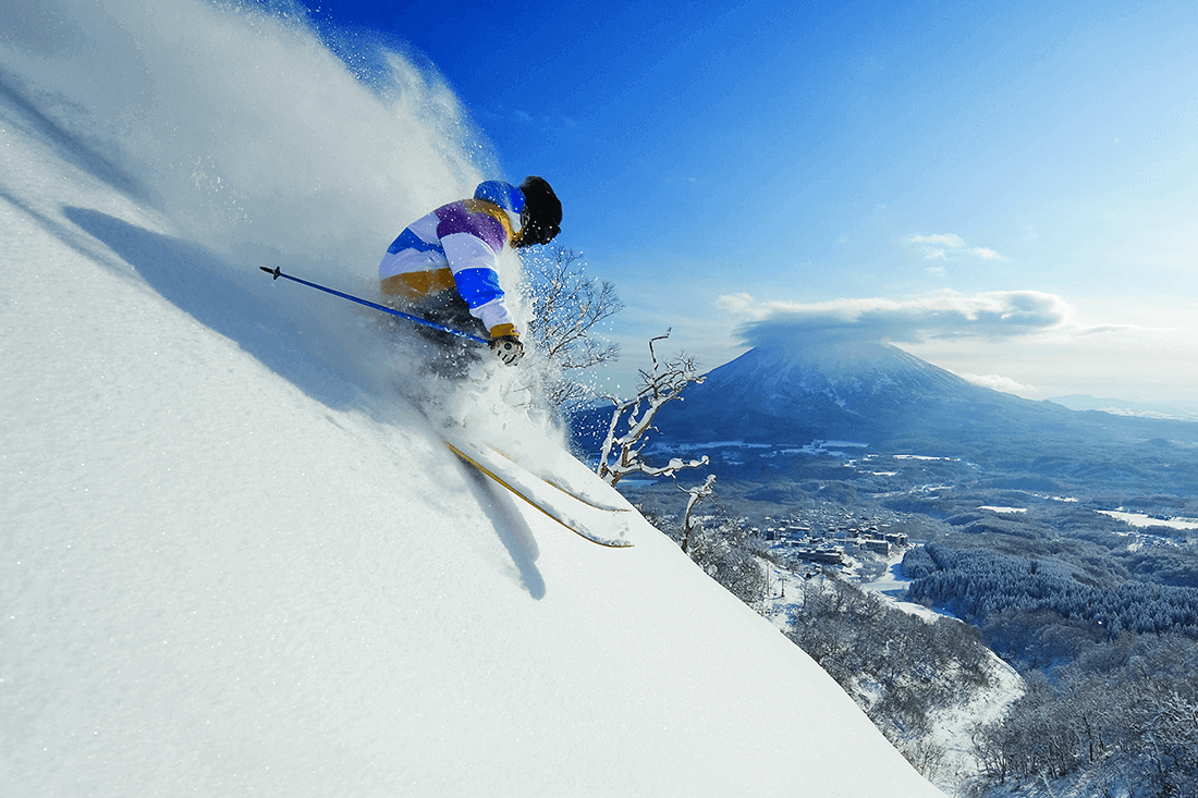 A skier creates a cloud of powder down a steep slope with a Japanese volcano in the background beneath a blue sky.