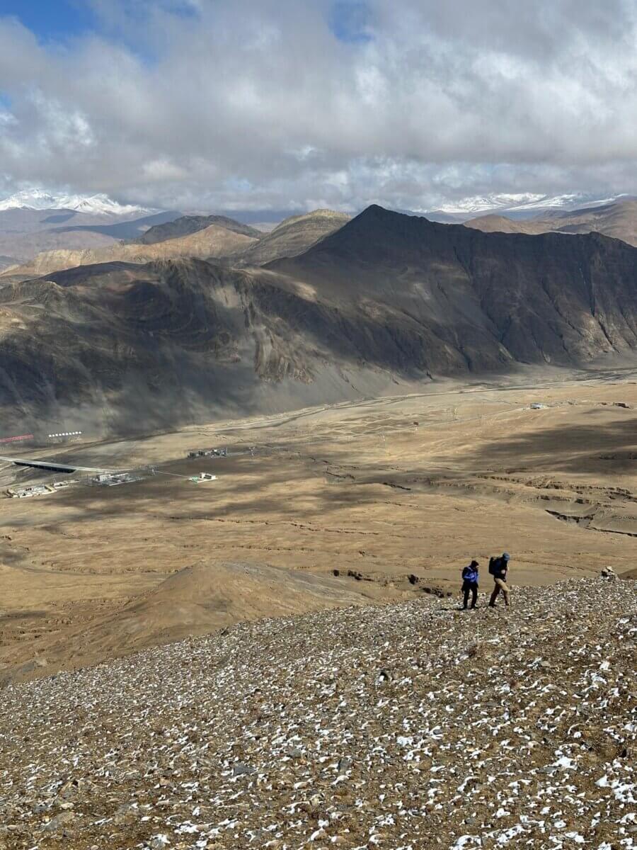 Climbers ascend a brown rocky hill with small mountains in the background on the Tibetan plateau.