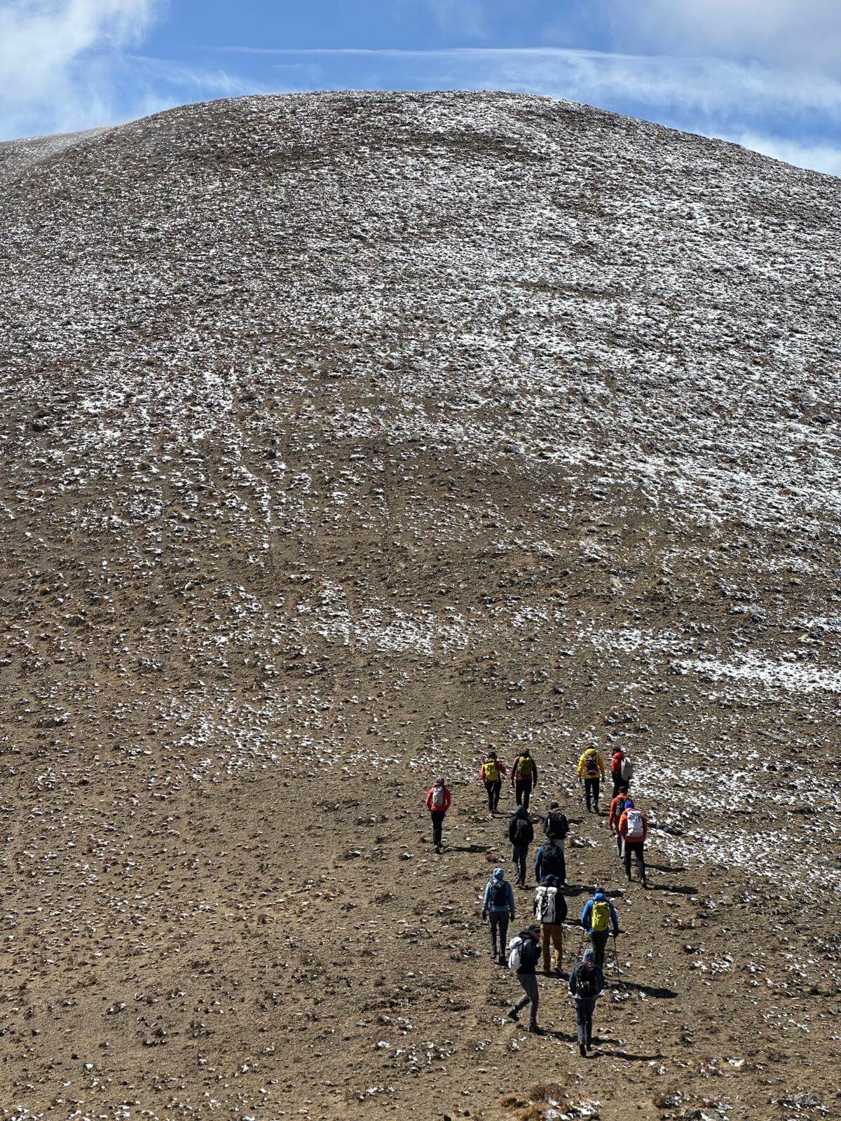 A group of climbers ascend a large, rocky hill that has a little snow on it while acclimatizing in Tingri, Tibet.