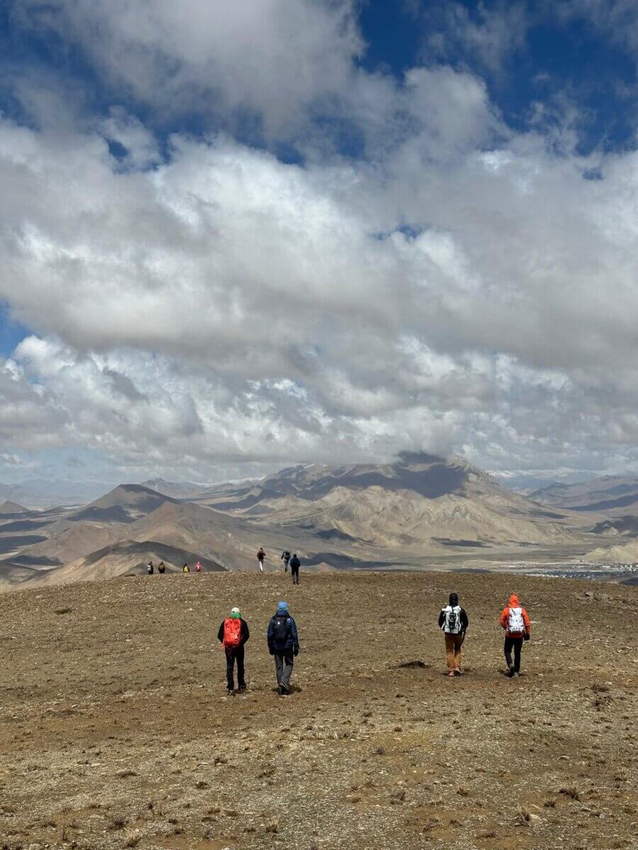 Climbers stand on a grassy flat looking out at the hilly Tibetan plateau beneath a cloudy sky.