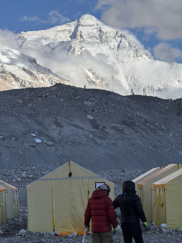 Two climbers in front of yellow living tents look up at the North Side of Mt. Everest beyond a shadowy hill.