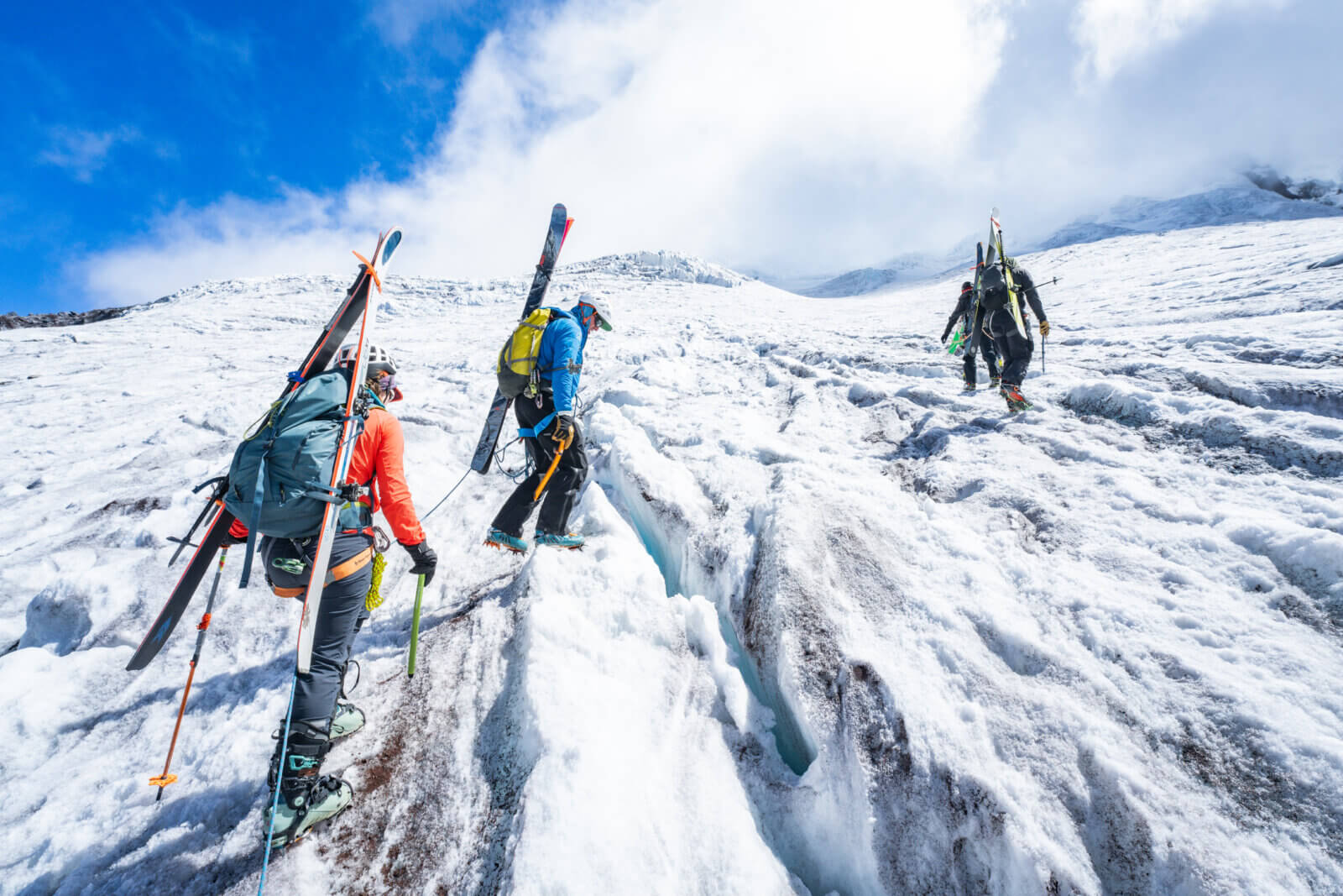 Three skiers step over small crevasses on a glacier beneath a blue cloudy sky during an international backcountry ski expedition with Alpenglow Expeditions..