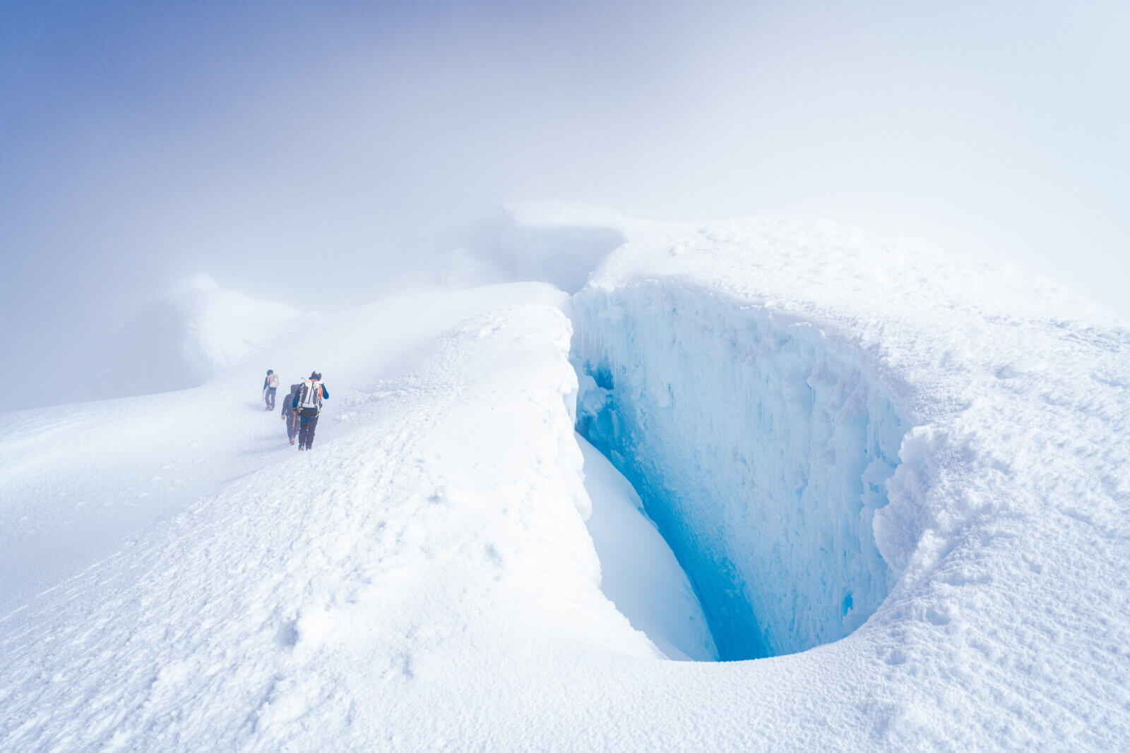 Three skiers descend to the left of a large, round crevasse with a blue crack at the bottom in misty conditions during an international backcountry ski expedition with Alpenglow Expeditions..