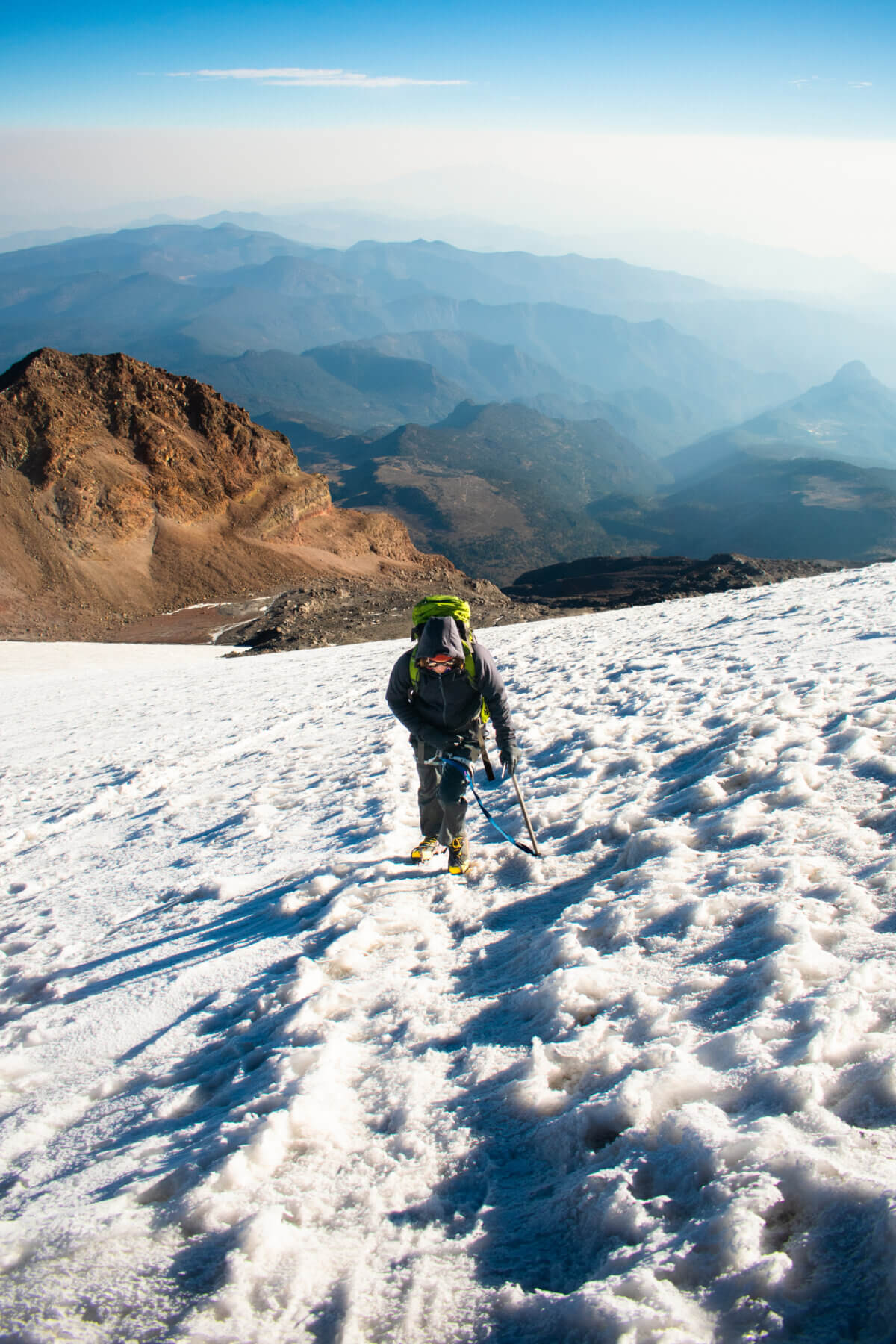 A climber ascends a snowy glacier with mountains in the background on Pico de Orizaba. 