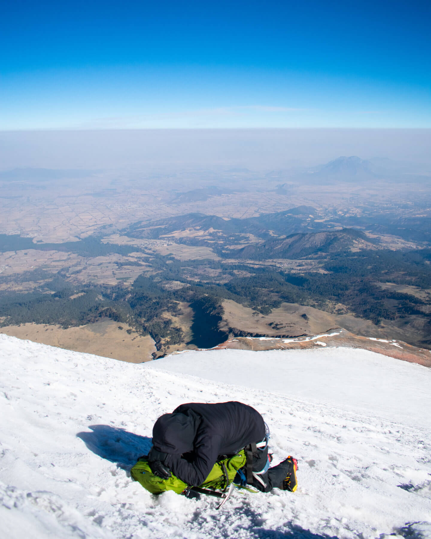 A climber in black lays over his green backpack on a white snow slope in front of a blue sky and a forested valley. 
