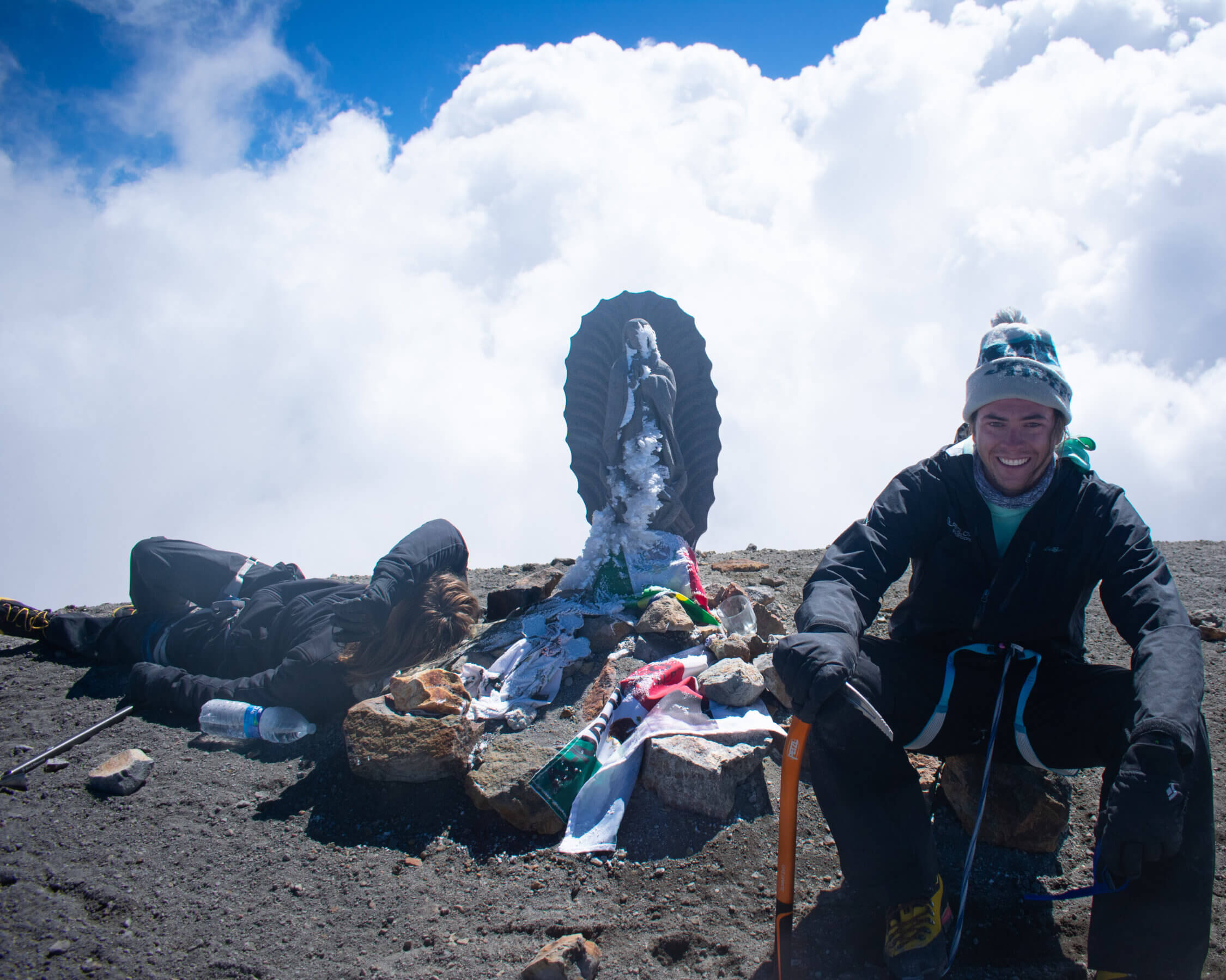 two climbers sit in front of puffy white clouds on the rocky gray summit of pico de orizaba with a small statue of the Virgin Mary wrapped in flags between them. 