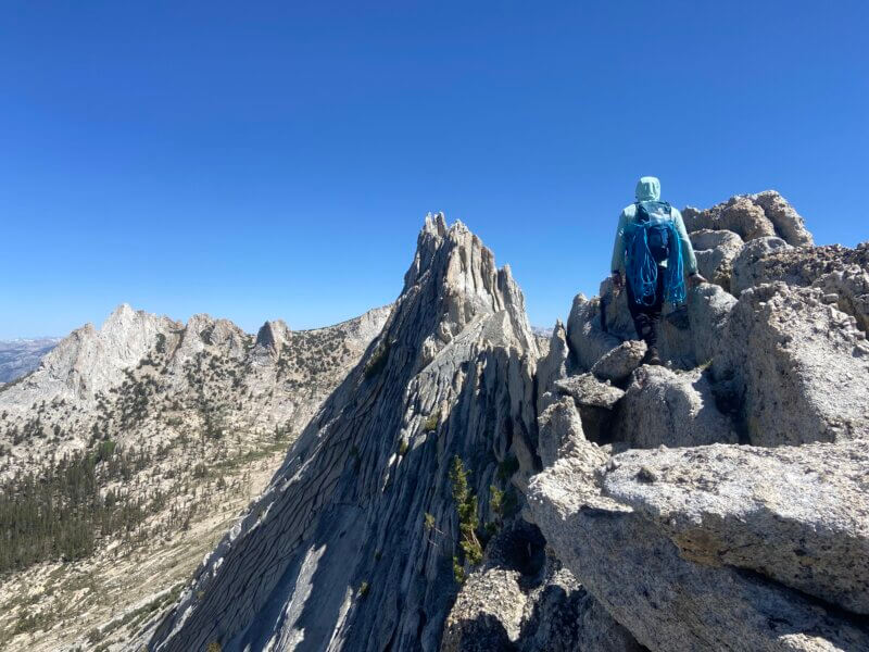 A climber with a blue rope and backpack walks along a sharp granite spine beneath a clear sky.