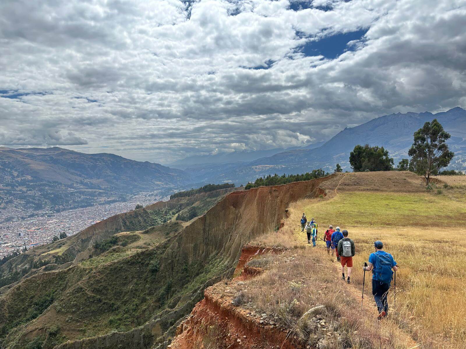 A group of people walk along a ridge with golden grass beneath a cloudy sky with obscured mountains in the background.