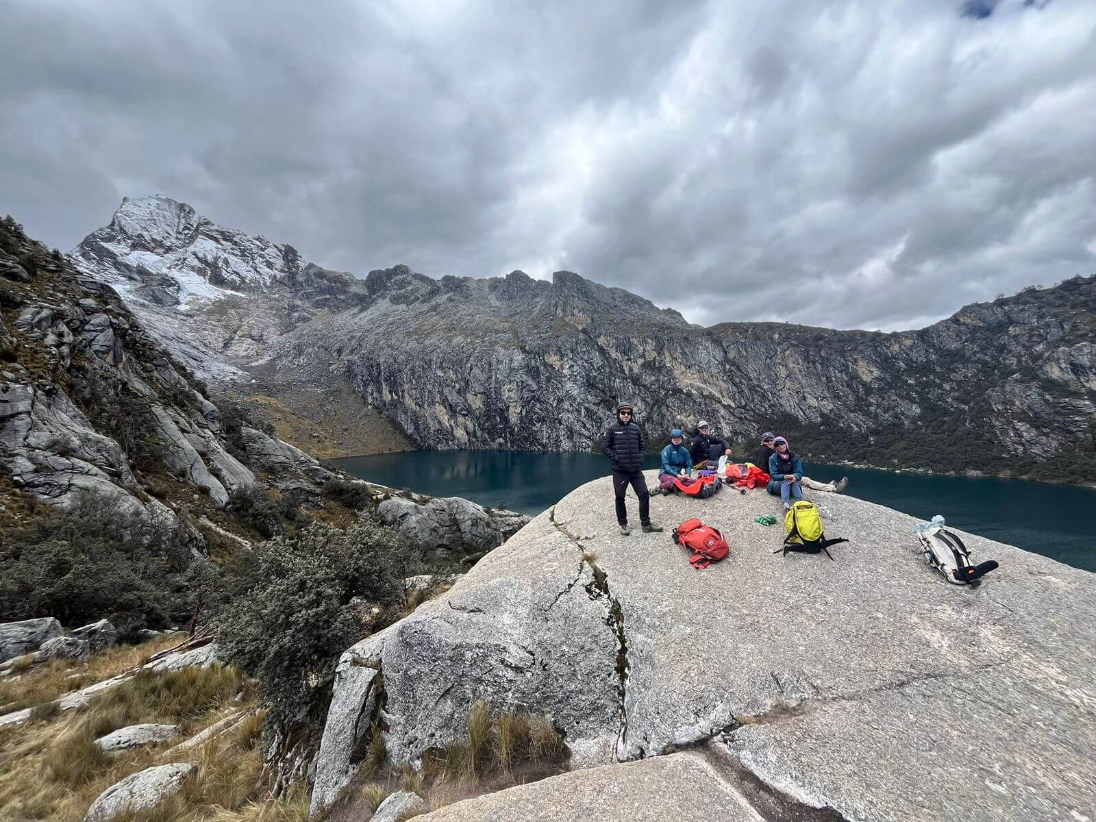 A group of hikers stands on a gray boulder positioned in front of a deep blue alpine lake surrounded by mountains beneath a cloudy sky.