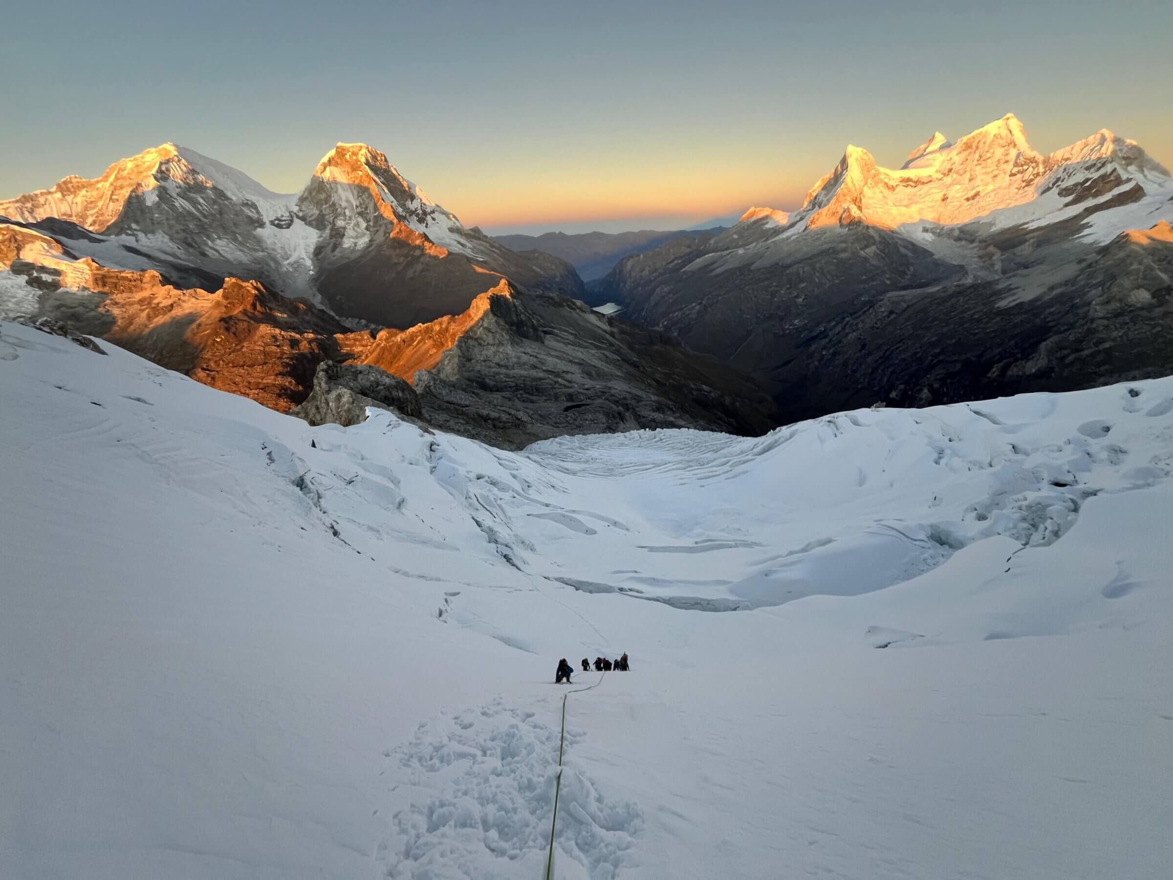 A group of climbers looks very small in the center of a wide snowfield with large mountains behind them in golden light.