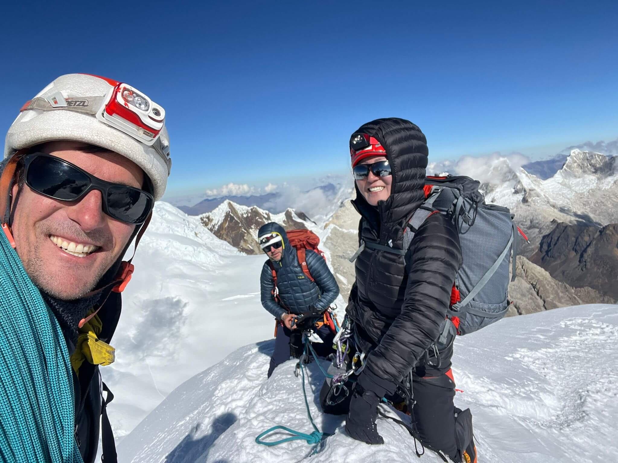 A selfie from a guide with a helmet and sunglasses and a rope with two clients dressed similarly on a snowy mountain ridge with clear blue skies. 
