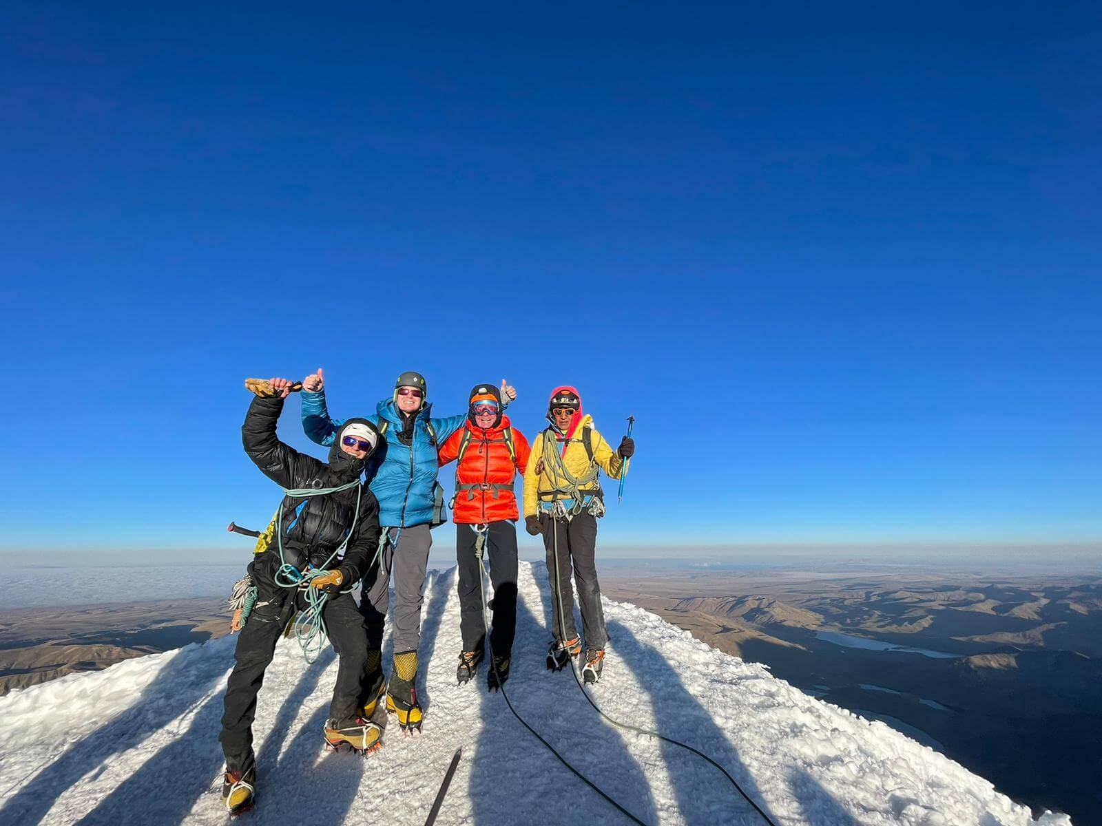 A group of climbers stand side by side on a snowy summit beneath a clear blue sky.