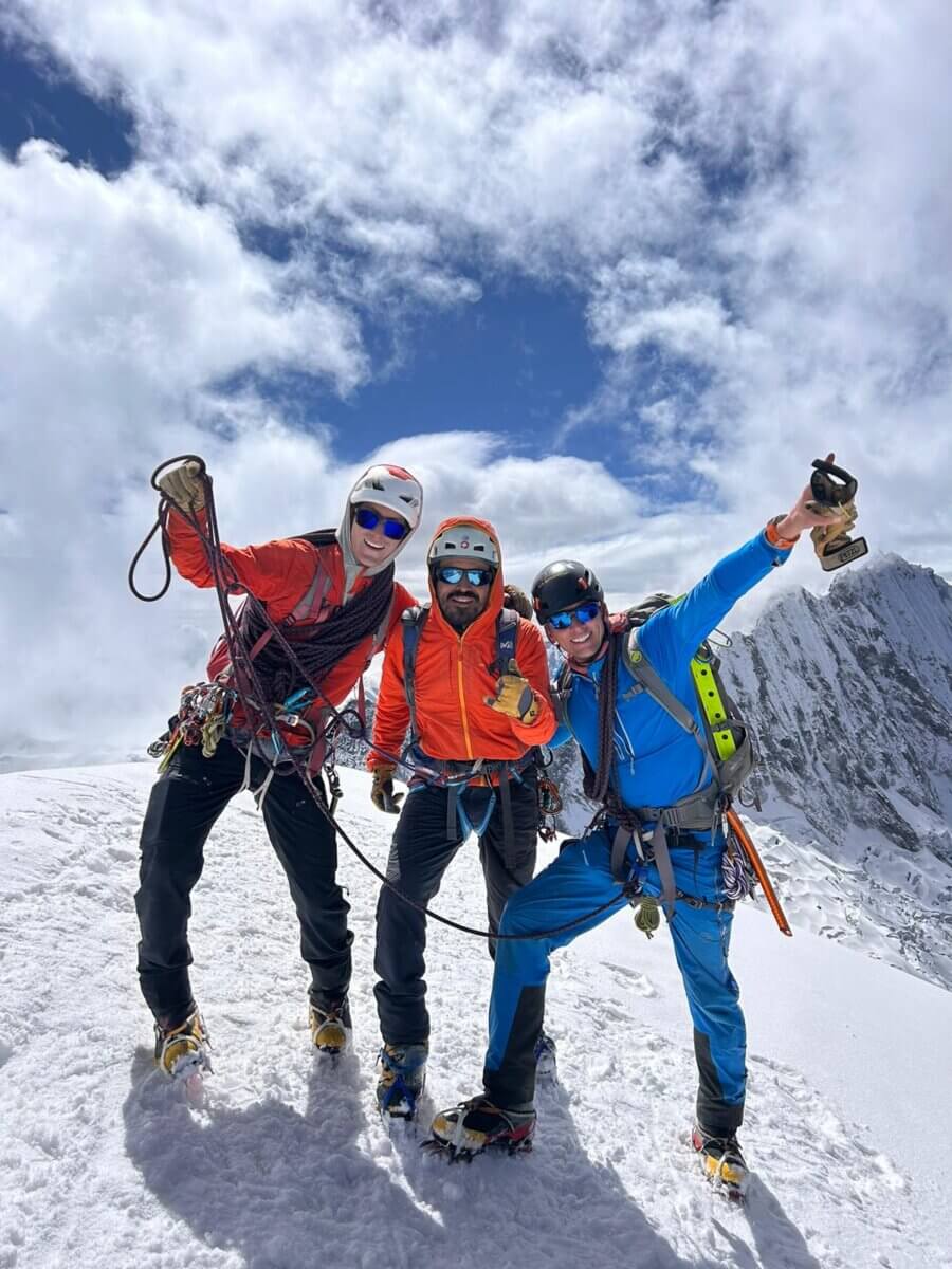 Three climbers stand with arms outstretched on a snowy mountain under a cloudy sky.