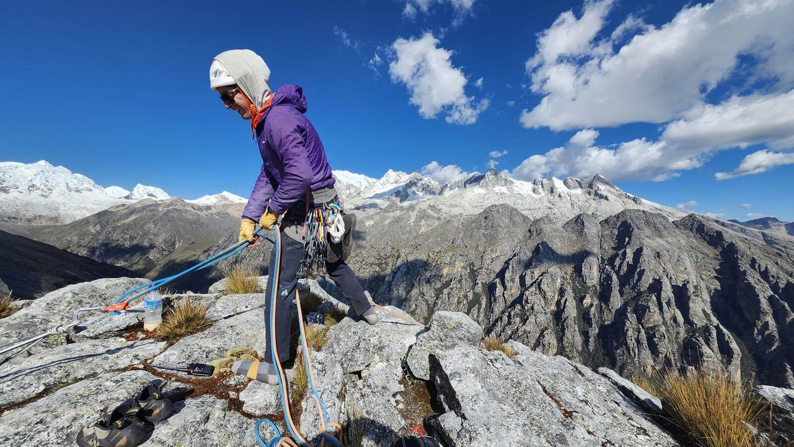 A mountain guide in a purple jacket manages ropes on a rocky mountaintop beneath a cloudy sky.