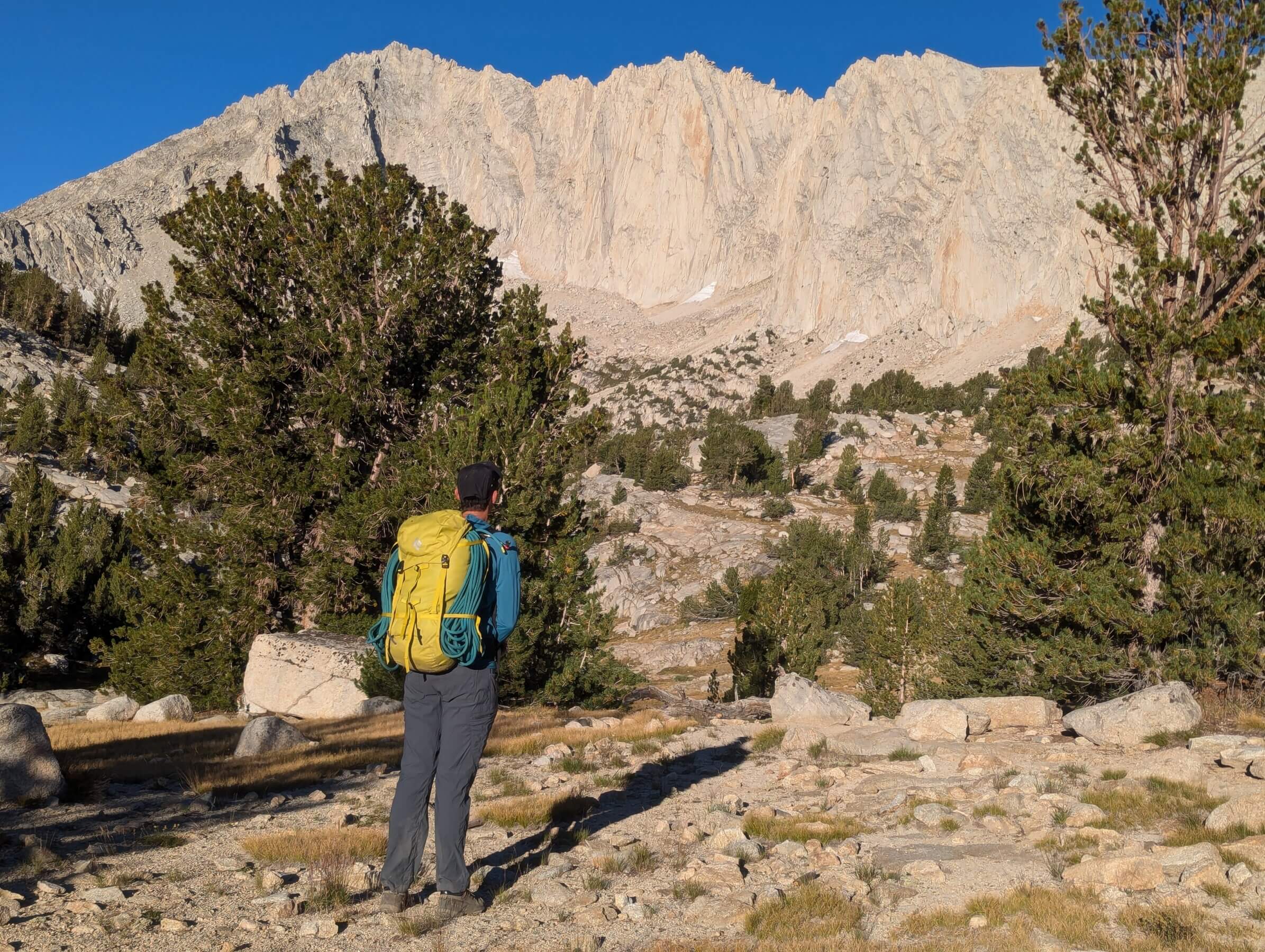 a climber in a blue jacket with a yellow pack stands in front of an alpine lake looking up at a white granite wall