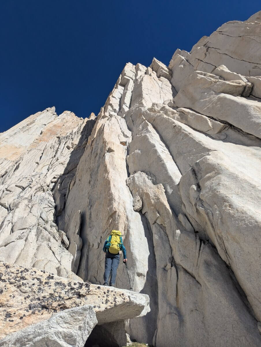 a climber with a yellow backpack stands beneath a steep white granite crack beneath a dark blue sky