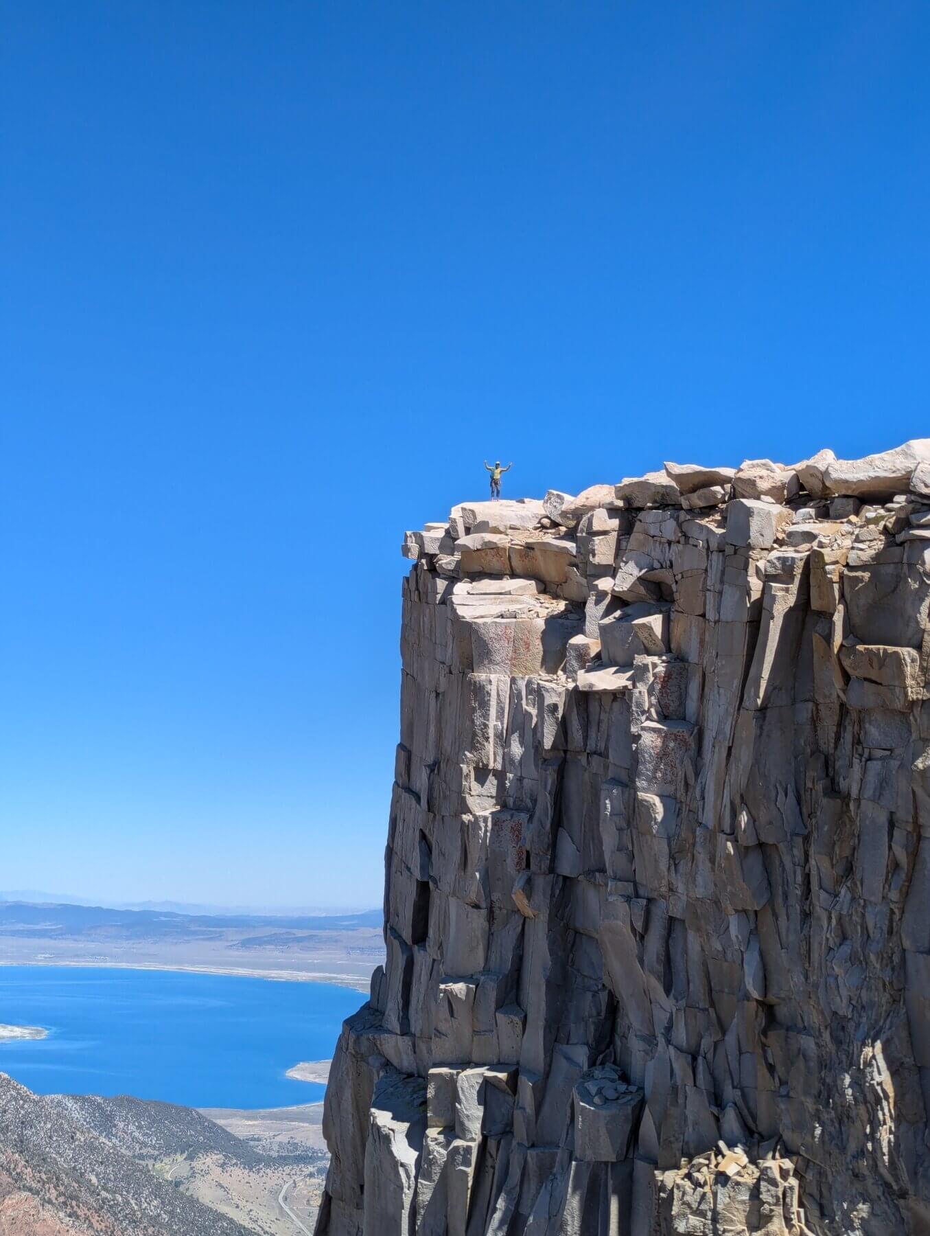 a rock climber stands atop a white pillar of rock with a blue lake far in the background