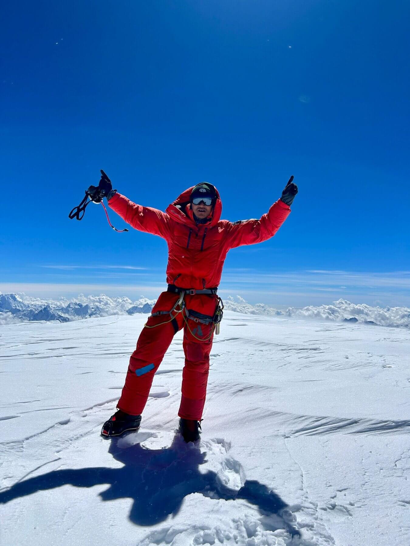 A Sherpa in a red down suit stands on the summit of Cho Oyu, a plain white snow field with a blue sky behind him. 