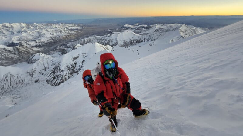 A climber in a red down suit climbs up a steep snow slope with the Himalaya behind at sunrise.